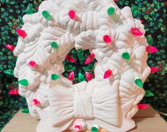 May include: A white ceramic Christmas wreath with a large bow at the bottom. The wreath is decorated with red and green lights. The wreath is on a wooden surface with a festive background.