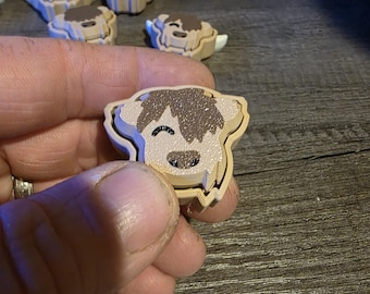 Op de afbeelding: Een close-up van een klein, houten, gelaagd, bruin en wit Highland-koeienbeeldje. Het beeldje heeft een glimlachend gezicht en is gemaakt van meerdere lagen hout.