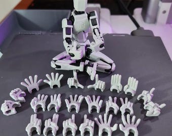Op de afbeelding: Een wit en grijs gearticuleerd figuur met meerdere verwisselbare handposities. Het figuur zit met verschillende handbevestigingen voor zich, die verschillende gebaren en posities laten zien. Het figuur heeft een blokkerig ontwerp.