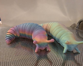 Op de afbeelding: Twee kleurrijke, beweegbare slakken. Eén is een regenboog van roze, paars en blauw, de andere een pastelmix van geel, groen en blauw. De speeltjes liggen op een zilveren, geperforeerd oppervlak.