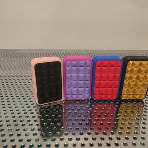 May include: Four rectangular, colorful objects with textured surfaces. The objects are pink, purple, blue, and gold. The pink object has a black surface, the purple has a purple surface, the blue has a red surface, and the gold has a gold surface.