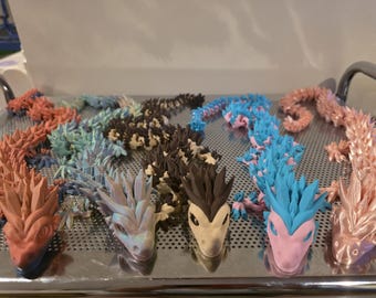 Op de afbeelding: Een verzameling kleurrijke, beweegbare drakenfiguren in verschillende tinten koraal, blauw, bruin en roze. De draken zijn gerangschikt op een metalen dienblad en tonen hun gedetailleerde, stekelige ontwerpen en unieke kleurencombinaties.
