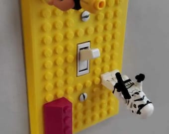 Op de afbeelding: Een gele Lego-thema lichtschakelaar cover met een witte schakelaar. De cover is versierd met Lego minifiguren, waaronder een zwart gekleed figuur en een Stormtrooper. Een rode Lego-steen is aan de onderkant bevestigd.