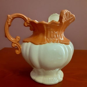 May include: A vintage-style ceramic pitcher with a cream-colored body and a pumpkin-orange rim and handle. The pitcher has a bulbous shape with a decorative handle and a ruffled rim. The handle has an ornate, scrolled design.