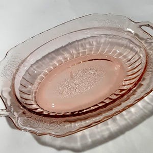 May include: A vintage pink glass serving dish with an oval shape. The dish has a scalloped edge and floral detailing around the rim. The interior features a raised oval section with a floral pattern. Two handles are on either side.