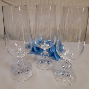 May include: Three clear champagne flutes with a crystal ball design on the stem. The flutes are tall and slender, with a wide bowl and a narrow base. Blue flowers are in the background.
