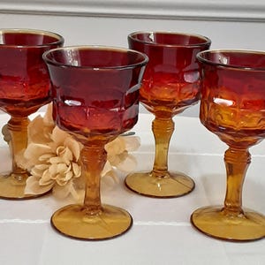 May include: Four vintage amber-colored wine glasses. Each glass features a textured, geometric pattern on the bowl and a stem with a round base. The glasses are arranged on a white surface, with a decorative flower in the background.