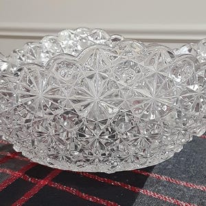 May include: A clear, decorative glass bowl with a starburst pattern. The bowl has a scalloped rim and sits on a red, black, and gray plaid surface. The bowl is likely used for serving.