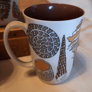 K&ouml;nnte beinhalten: Wei&szlig;e Keramiktasse mit braunem Innenraum und gebogenem Henkel. Die Tasse ist mit verschiedenen Muscheldarstellungen in Braun-, Beige- und Graut&ouml;nen verziert. Das Design umfasst einen Spiral-Nautilus, eine Kegelmuschel und andere Meeresmotive.