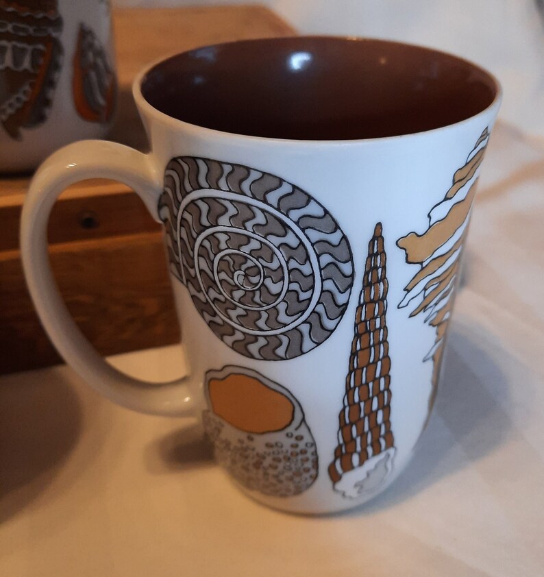 K&ouml;nnte beinhalten: Wei&szlig;e Keramiktasse mit braunem Innenraum und gebogenem Henkel. Die Tasse ist mit verschiedenen Muscheldarstellungen in Braun-, Beige- und Graut&ouml;nen verziert. Das Design umfasst einen Spiral-Nautilus, eine Kegelmuschel und andere Meeresmotive.