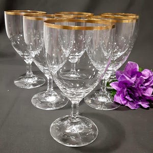 May include: Six clear wine glasses with gold rims are arranged on a dark surface. The glasses have a classic design with a rounded bowl and a slender stem. A purple flower is in the background.