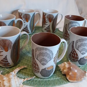 K&ouml;nnte beinhalten: Eine Reihe wei&szlig;er Keramikbecher mit Muscheldesign in Orange, Grau und Braun. Die Becher haben eine braune Innenseite und wei&szlig;e Henkel. Eine Muschel ist im Vordergrund zu sehen. Die Becher stehen auf einer gr&uuml;nen Unterlage.