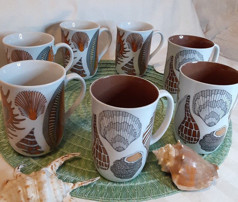 K&ouml;nnte beinhalten: Eine Reihe wei&szlig;er Keramikbecher mit Muscheldesign in Orange, Grau und Braun. Die Becher haben eine braune Innenseite und wei&szlig;e Henkel. Eine Muschel ist im Vordergrund zu sehen. Die Becher stehen auf einer gr&uuml;nen Unterlage.