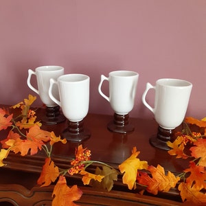 May include: Four white coffee mugs with brown bases are arranged on a dark wooden surface. The mugs have a classic design with curved handles. A garland of orange and yellow autumn leaves and berries decorates the scene, suggesting a fall theme.