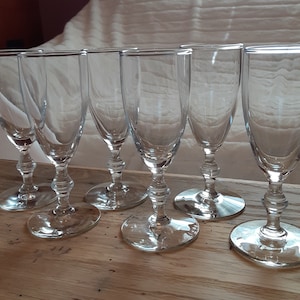May include: Six clear, stemmed glasses with rounded bowls and decorative stems. The glasses sit on a wooden surface, with a white background. The glasses are empty and appear to be made of glass.