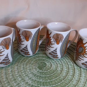 K&ouml;nnte beinhalten: Vier wei&szlig;e Keramikbecher mit Muscheldesign in Orange, Grau und Braun. Jeder Becher hat einen Henkel und ein sich wiederholendes Muster aus Muscheln und anderen Meereselementen. Die Becher stehen auf einem gr&uuml;nen Platzset.