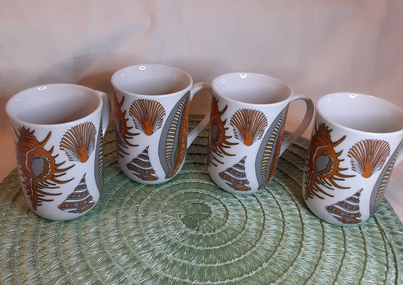 K&ouml;nnte beinhalten: Vier wei&szlig;e Keramikbecher mit Muscheldesign in Orange, Grau und Braun. Jeder Becher hat einen Henkel und ein sich wiederholendes Muster aus Muscheln und anderen Meereselementen. Die Becher stehen auf einem gr&uuml;nen Platzset.