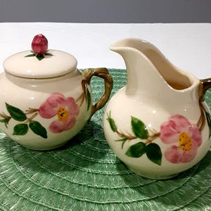 May include: Cream-colored ceramic sugar bowl with lid and creamer set. Both pieces are decorated with pink rose floral designs and brown twig-like handles. The sugar bowl has a red bud finial.