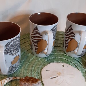 K&ouml;nnte beinhalten: Drei wei&szlig;e Keramikbecher mit braunem Innenraum und wei&szlig;en Griffen. Jeder Becher hat ein Muscheldesign in Braun, Beige und Grau. Die Becher sind auf einer gr&uuml;nen Matte mit Muscheln und anderen dekorativen Gegenst&auml;nden angeordnet.