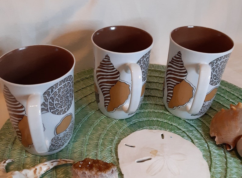 K&ouml;nnte beinhalten: Drei wei&szlig;e Keramikbecher mit braunem Innenraum und wei&szlig;en Griffen. Jeder Becher hat ein Muscheldesign in Braun, Beige und Grau. Die Becher sind auf einer gr&uuml;nen Matte mit Muscheln und anderen dekorativen Gegenst&auml;nden angeordnet.