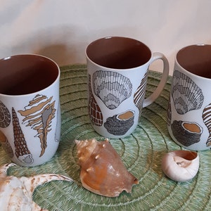 K&ouml;nnte beinhalten: Drei wei&szlig;e Keramikbecher mit braunen Innenr&auml;umen und Griffen. Jeder Becher ist mit verschiedenen Muscheldesign in Braun-, Grau- und Beiget&ouml;nen verziert. Muscheln und eine gr&uuml;ne Tischmatte sind ebenfalls sichtbar.
