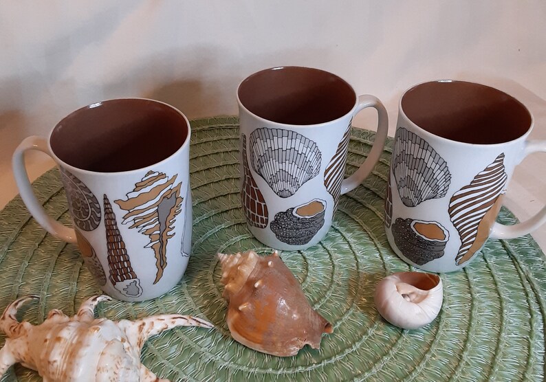K&ouml;nnte beinhalten: Drei wei&szlig;e Keramikbecher mit braunen Innenr&auml;umen und Griffen. Jeder Becher ist mit verschiedenen Muscheldesign in Braun-, Grau- und Beiget&ouml;nen verziert. Muscheln und eine gr&uuml;ne Tischmatte sind ebenfalls sichtbar.