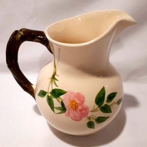 May include: Cream-colored ceramic pitcher with a brown handle designed to look like a branch. The pitcher is decorated with a pink rose and green leaves. The pitcher has a curved spout.