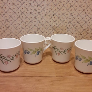 May include: Four white ceramic mugs with floral designs. Each mug features a handle and is decorated with a pattern of pink and blue flowers with green leaves. The mugs are arranged on a wooden surface.