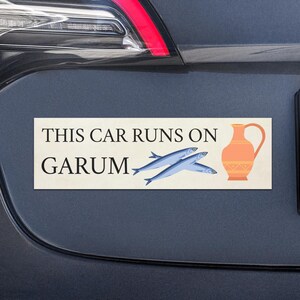 K&ouml;nnte beinhalten: Rechteckiger Aufkleber auf einem dunkelgrauen Auto. Der Aufkleber lautet "THIS CAR RUNS ON GARUM" mit Illustrationen von zwei blauen Fischen und einer orangefarbenen Amphore.