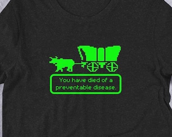 Puede incluir: Camiseta gris oscuro con una imagen pixelada verde de una carreta cubierta tirada por un buey. El texto "You have died of a preventable disease." está debajo de la imagen. La camiseta se muestra con vaqueros y accesorios.