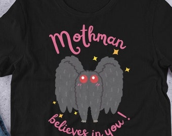 Puede incluir: Camiseta negra con un diseño de Mothman de dibujos animados. El Mothman tiene ojos rojos y alas grises, con el texto "Mothman believes in you!" en rosa. La camiseta se muestra sobre una superficie plana.
