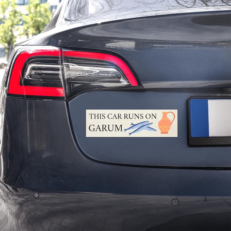 K&ouml;nnte beinhalten: Rechteckiger Aufkleber auf einem dunkelgrauen Auto. Der Aufkleber lautet "THIS CAR RUNS ON GARUM" mit Illustrationen von zwei blauen Fischen und einer orangefarbenen Amphore.