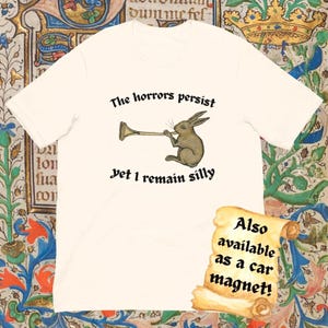Puede incluir: Camiseta color crema con una ilustraci&oacute;n de un conejo marr&oacute;n tocando una trompeta, con el texto "The horrors persist, yet I remain silly." Un pergamino indica que el dise&ntilde;o tambi&eacute;n est&aacute; disponible como im&aacute;n para coche.