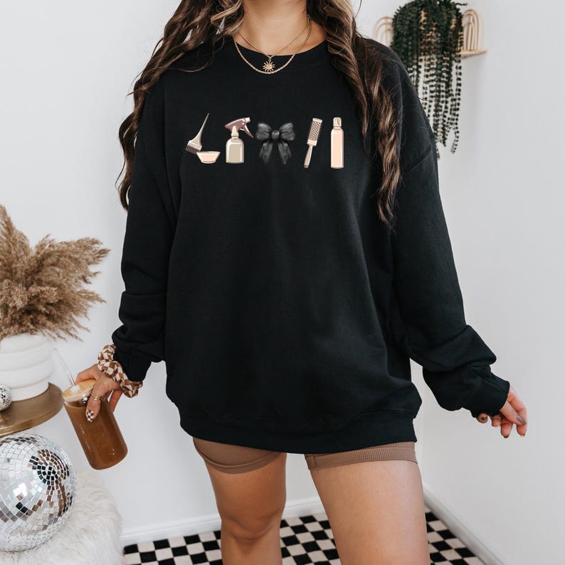 May include: Black sweatshirt with a graphic of hair styling tools, including a spray bottle, hair dye, a bow, a comb, and a bottle of hair product.