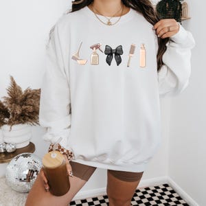 May include: White sweatshirt with a graphic of hair styling tools, including a brush, spray bottle, bowl, black bow, and hairspray bottle.