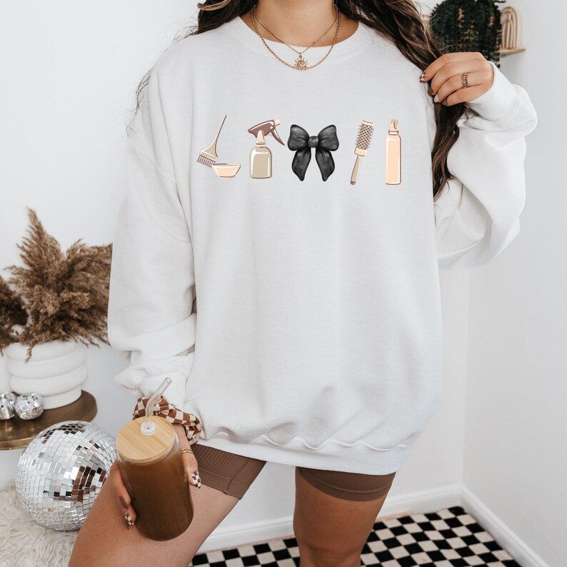 May include: White sweatshirt with a graphic of hair styling tools, including a brush, spray bottle, bowl, black bow, and hairspray bottle.
