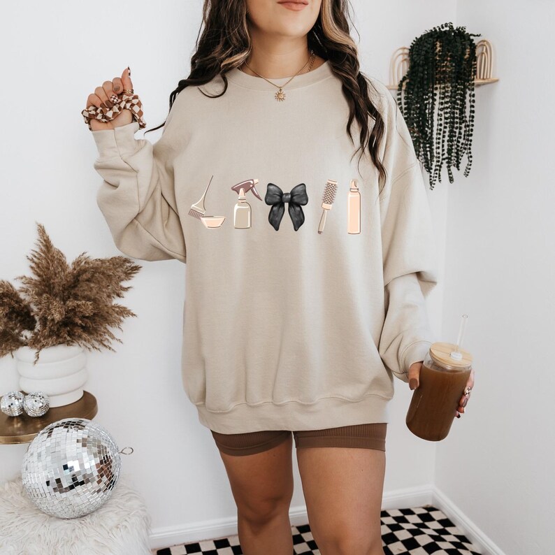 May include: A beige sweatshirt with a graphic of hair styling tools, including a spray bottle, a bowl, a hairbrush, a black bow, and a bottle of hair dye. The sweatshirt is being worn by a person.