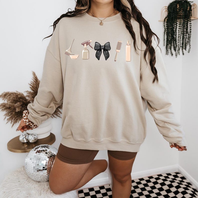 May include: A beige sweatshirt with a graphic design of hair styling tools, including a spray bottle, a bowl, a hairbrush, a black bow, and a bottle of hair product.