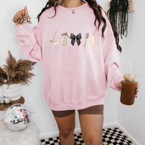 May include: A pink sweatshirt with a graphic of hair styling tools, including a spray bottle, a bowl, a hairbrush, a black bow, and a bottle of hair dye. The sweatshirt is being worn by a person.