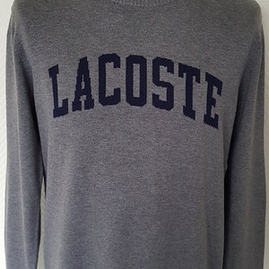 Könnte beinhalten: Ein grauer Langarm-Pullover mit einem dunkelblauen "LACOSTE"-Logo auf der Vorderseite.