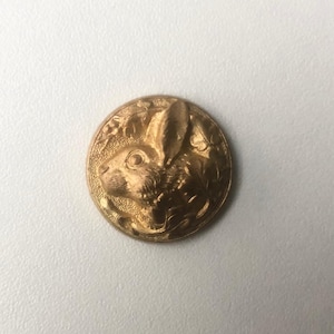 May include: A gold-coloured button featuring a detailed relief of a rabbit's head in profile, encircled by foliage. The button is round with a slightly textured surface, giving it a vintage look. The rabbit's features are clearly defined.