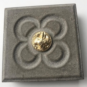 Puede incluir: Una baldosa cuadrada de hormigón gris con un diseño de flor de cuatro pétalos en relieve negativo. Centrado en la flor hay un botón metálico circular de tono dorado con el perfil detallado de un conejo rodeado de motivos vegetales.