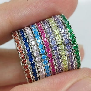 May include: A collection of colorful, stackable rings. Each ring is adorned with a row of small, vibrant gemstones in a rainbow of colors, including red, orange, blue, pink, green, and purple. The rings are held in a hand.