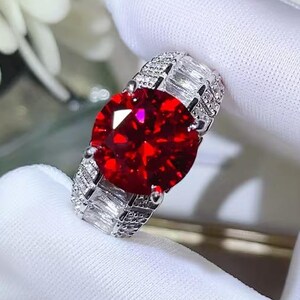 Op de afbeelding: Een zilveren ring met een grote, ronde, helderrode edelsteen als middelpunt. De band is bezet met kleinere, rechthoekige, heldere edelstenen. De ring wordt getoond op een witte ondergrond, wat de schittering benadrukt.