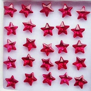 Op de afbeelding: Een verzameling kleine, facetgeslepen, stervormige edelstenen in een levendige rode kleur. De edelstenen zijn in een raster gerangschikt in een witte, vierkante container. Geschikt voor het maken van sieraden of knutselprojecten.