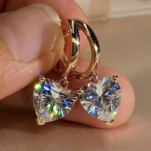 Könnte beinhalten: Ein Paar goldfarbene Creolen mit herzförmigen, klaren, facettierten Edelsteinen. Die Edelsteine reflektieren einen Regenbogen an Farben. Die Ohrringe werden von den Fingern einer Person gehalten.