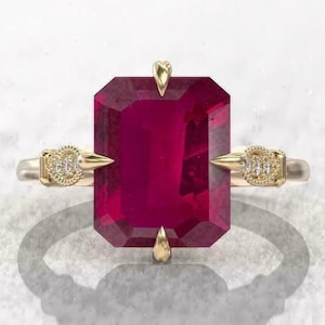 Op de afbeelding: Een gouden ring met een grote, rechthoekige, dieprode edelsteen. De ring heeft een delicaat ontwerp met kleine diamantaccenten aan weerszijden van de edelsteen. De ringband is van gepolijst goud.