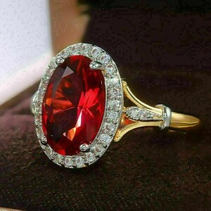 Op de afbeelding: Een gouden ring met een grote, ovaalvormige, rode edelsteen omringd door een halo van kleine, heldere stenen. De ringband is goudkleurig met zilveren details, wat een klassiek en elegant ontwerp laat zien.