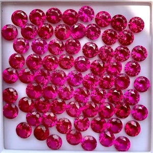 Può includere: Una collezione di gemme rotonde, sfaccettate, rosa acceso. Le gemme sono disposte in un contenitore quadrato bianco. Le pietre hanno un taglio brillante, che riflette la luce e crea un effetto scintillante. L'impressione generale è di lusso e valore.