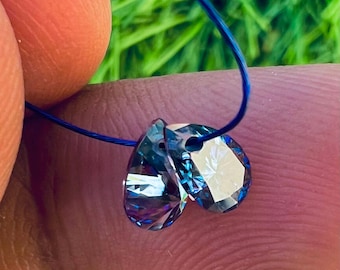 May include: Two iridescent teardrop-shaped crystal beads, each with multiple facets, are strung on a thin blue cord. The crystals reflect a spectrum of colors, including blue, purple, and green. The background is blurred, with green grass visible.